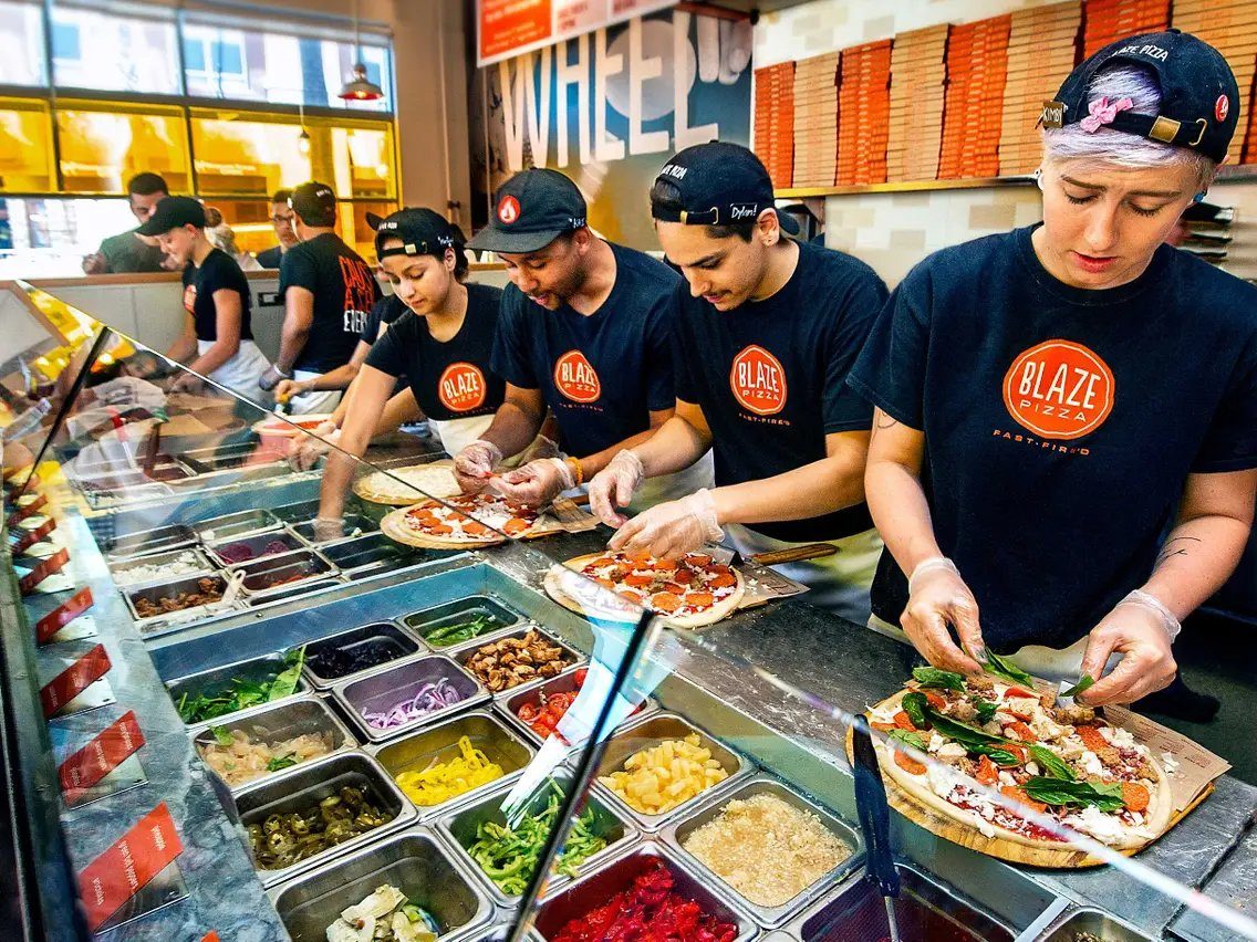 A team building pizzas side by side along a line of topping wells