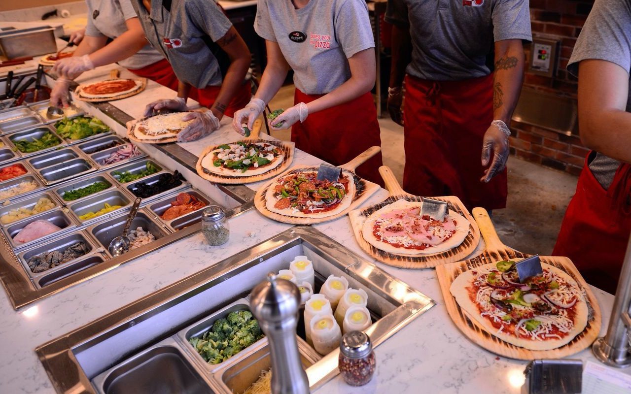 Looking down on a make-line: wells of vegetables, cheeses and meats on one side, pizzas being built on wooden peels on the other